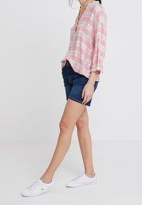 Vaaleanpunainen ja valkoinen ruutukuosisekasvi, jossa on V-pääntie ja kolmannes hihat, yhdistettynä tummiin farkkushortseihin ja valkoisiin lenkkareihin.