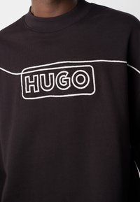 Sweat-shirt noir à col rond avec logo "HUGO" en contour blanc et motif de lignes reliées sur la poitrine, porté par une personne.