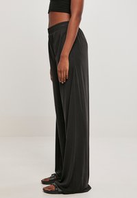 Person trägt weite schwarze Hose und schwarze Sandalen, steht seitlich vor einfachem hellem Hintergrund.