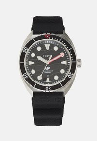 Montre avec un bracelet en silicone noir, un boîtier en acier inoxydable, un cadran texturé noir, des index lumineux, une aiguille des secondes rouge et une lunette tournante.