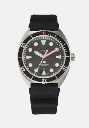 Montre avec un bracelet en silicone noir, un boîtier en acier inoxydable, un cadran texturé noir, des index lumineux, une aiguille des secondes rouge et une lunette tournante.