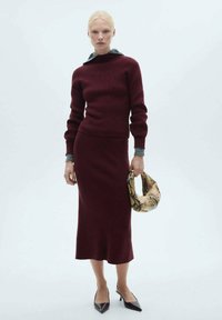 Burgunderfarbener, gestrickter Pullover und passender Rock mit einer taillierten Silhouette. Grauer Kapuzenakzent; hält eine gemusterte, strukturierte Handtasche. Hochhackige Schuhe.
