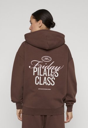 Kvinde med mørkt hår samlet i en knold iført en brun hættetrøje med teksten "Friday Pilates Class" i hvidt på ryggen, stående foran en ensfarvet baggrund.