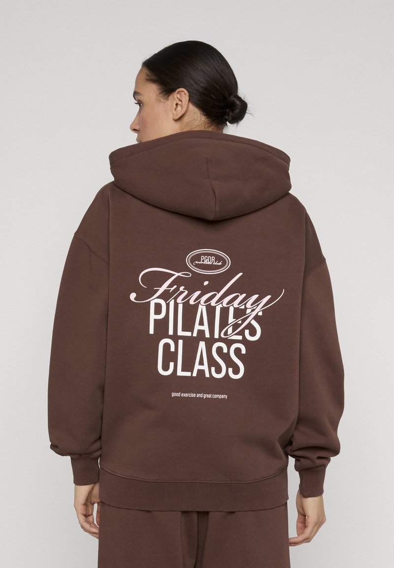 Femme aux cheveux foncés en chignon portant un sweat à capuche marron avec le texte "Friday Pilates Class" en blanc au dos, debout devant un fond uni.