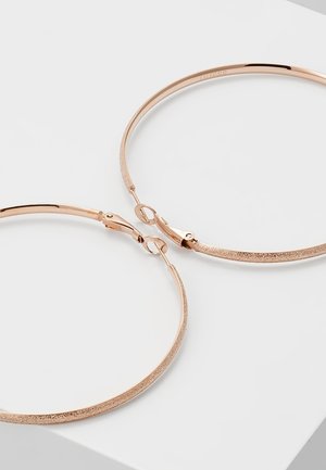 Boucles d’oreilles créoles fines en or rose avec surface texturée et fermoirs à charnière sur une surface blanche.