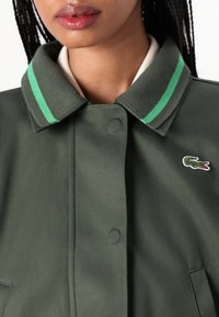 Groene jas met een contrasterende kraag met groene strepen, drukknoopjes en een klein geborduurd logo op de borst. Gladde stofstructuur.