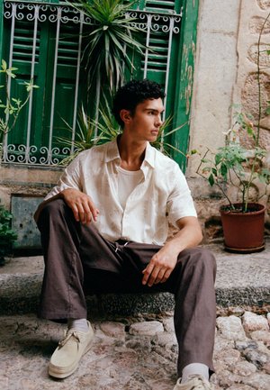 Junger Mann in weißem Hemd und brauner Hose sitzt auf einer Steintreppe, umgeben von grünen Pflanzen und mit einem grünen Fensterladen hinter sich.