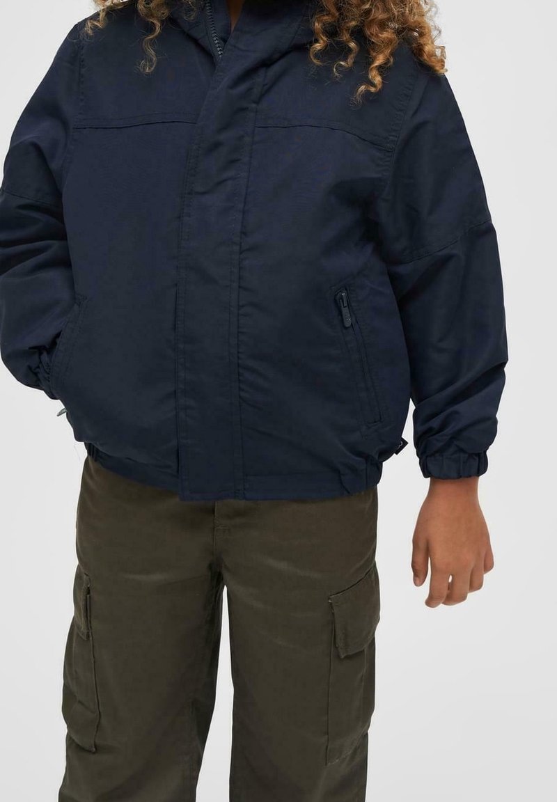 Enfant aux cheveux bouclés portant une veste zippée bleu marine et un pantalon cargo vert olive, debout devant un fond uni.