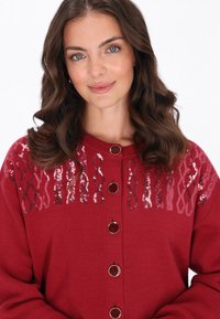 Cardigan roșu cu un model texturat, accentuat cu paiete la guler. Prezintă butoane circulare și mâneci lungi. Material moale.