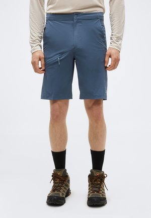 Férfi, kék cipzáras túranadrágot, bézs hosszú ujjú felsőt, fekete zoknit és barna túracipőt visel, fehér háttér előtt áll.