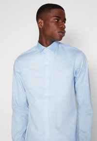Camisa de manga larga azul claro con cuello de botón, hecha de una tela suave que presenta una textura lisa y un frente con botones clásico.