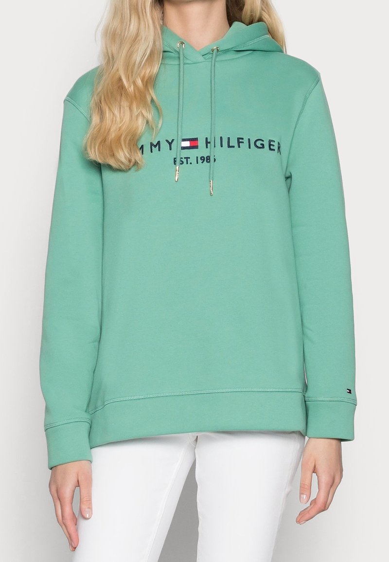 Personne portant un sweat à capuche Tommy Hilfiger vert clair avec logo et un pantalon blanc, debout les bras détendus le long du corps.