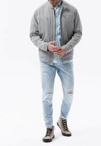 Grijze bomberjack met ritszakken, gelaagd over een lichtblauw denimhemd; gecombineerd met versleten lichtblauwe jeans en beige hoge laarzen.