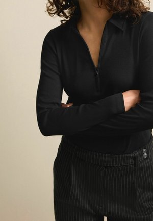 Kobieta o kręconych włosach ubrana w czarny, prążkowany, długi sweter z częściowym zamkiem błyskawicznym oraz czarne spodnie w prążki, ze skrzyżowanymi ramionami.