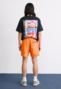 Svart kortermet t-skjorte med fargerik grafikk på baksiden. Oransje elastiske shorts med lommer. Modellen har på seg hvite sokker og grå joggesko.