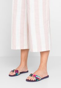 Lila platta sandaler med en knuten blommig rem i livliga färger, bärs med ljusrosa och vita randiga vida byxor.