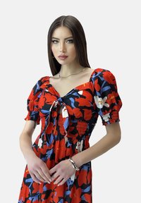 Vestito floreale con motivi rossi, blu e bianchi, maniche a sbuffo corte e chiusura con lacci frontale; arricciatura testurizzata in vita.