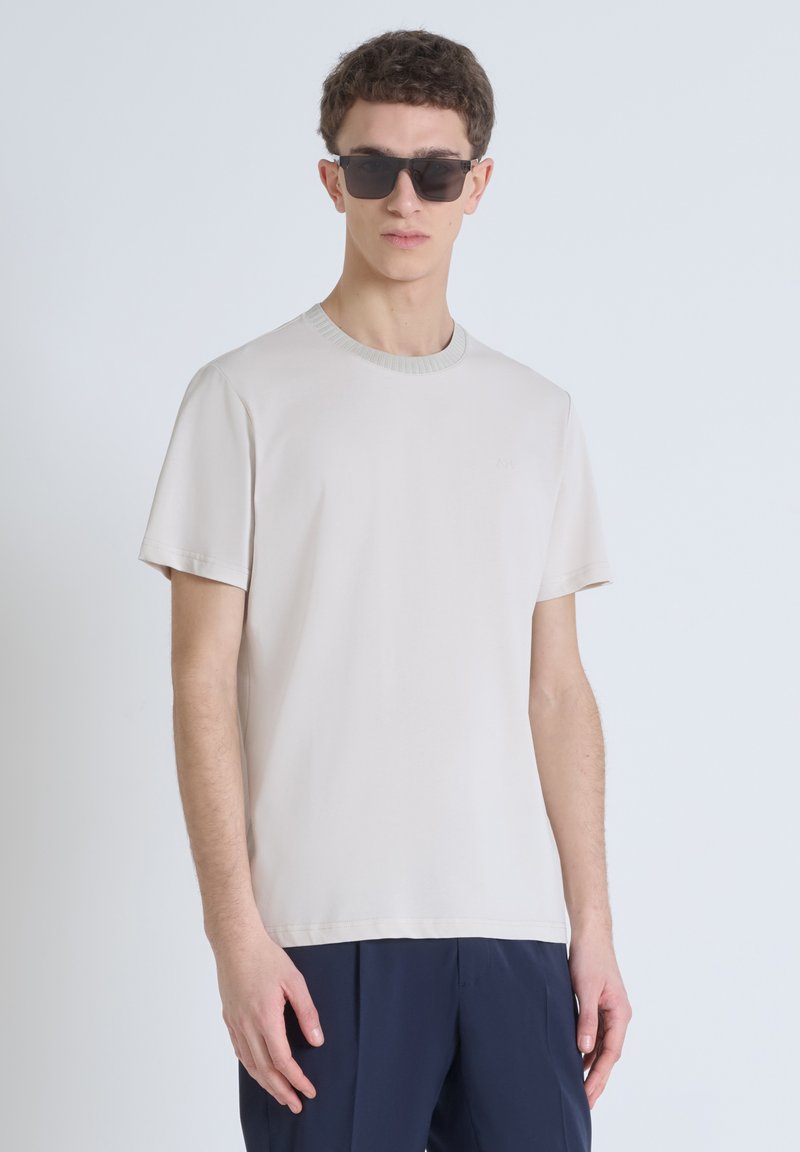 Camiseta de cuello redondo en color beige claro, hecha de algodón suave, con mangas cortas y un cuello texturizado, combinada con pantalones oscuros de color navy y gafas de sol.