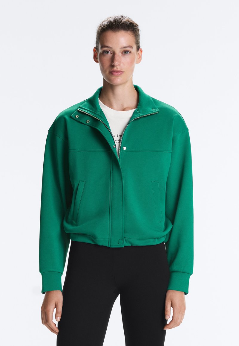 Femme portant une veste zippée verte par-dessus une chemise blanche et un pantalon noir, debout devant un fond blanc uni.