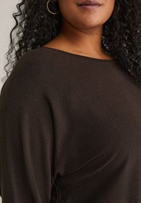 Hnědý svetr s volným střihem a širokým výstřihem, vyrobený z měkké tkaniny. Má texturovaný pletený vzor a krátké rukávy. Viditelné velké kruhové náušnice.