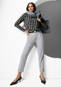 Zwart en grijs houndstooth-trui, grijze op maat gemaakte broek met geplooide manchetten, zwarte puntige hakken, grijze sjaal nonchalant over de schouder, zwarte riem.