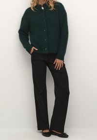 Cardigan verde scuro, dalla texture lavorata, con abbottonatura frontale e maniche lunghe, abbinato a pantaloni neri a vestibilità ampia. Mocassini neri completano l'outfit.