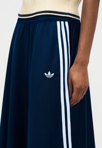 Jupe Adidas bleu marine avec trois bandes blanches et logo, portée avec un haut côtelé beige, montrant un gros plan de la taille et de la main.