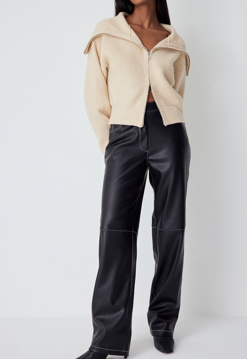 Femme portant un pull crème côtelé zippé avec un large col et un pantalon noir en similicuir taille haute avec coutures visibles.