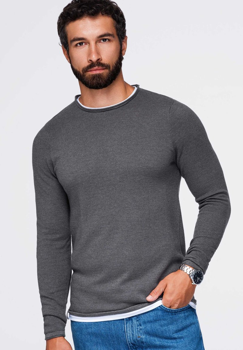 Homme avec une barbe portant une chemise grise texturée à manches longues, un jean bleu et une montre-bracelet argentée, debout avec une main dans la poche.