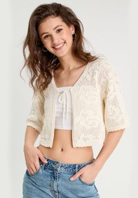 Cardigan en tricot crème à manches courtes, avec une ouverture frontale et un motif floral. Associé à un crop top blanc et à un short en denim taille haute.