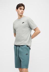 Νεαρός άνδρας που φοράει γκρι μπλουζάκι Nike και βερμούδα με κορδόνι Nike σε τυρκουάζ χρώμα, στέκεται με τα χέρια πίσω από την πλάτη, κοιτάζοντας προς τα πλάγια.