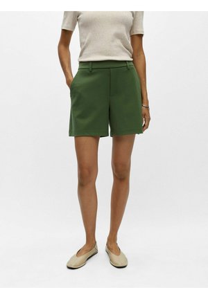 Kvinde iført grønne skræddersyede shorts, beige kortærmet top og beige flade sko, stående foran en ensfarvet baggrund.