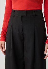 Pantaloni negri cu talie înaltă, cu pliuri și buzunare laterale, dintr-un material neted. Asortați cu un top lung cu mâneci lungi, texturat, de culoare roșie.