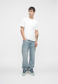 Młody mężczyzna stoi z rękami w kieszeniach, ubrany w białą koszulkę, jasnoniebieskie dżinsy, sneakersy oraz z widocznym tatuażem na lewym przedramieniu, na jednolitym tle.