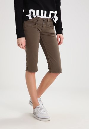 Person iført brune knælange shorts, en sort sweatshirt med delvist synlig hvid tekst og hvide sneakers, der står med benene krydset.