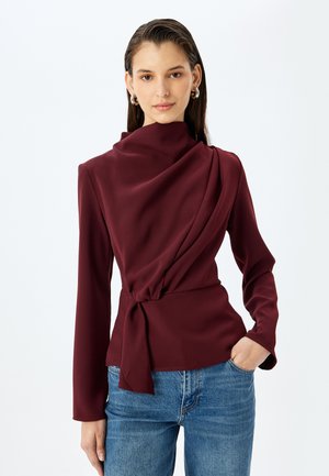 Bordowa górna część z drapowanym dekoltem, długimi rękawami i wiązaniem w talii, wykonana z gładkiego materiału; noszona z niebieskimi dżinsami.
