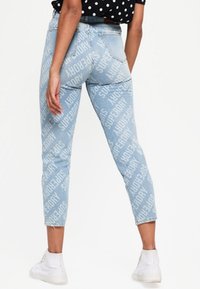 Jean en denim bleu clair avec un motif de logo "Superdry" sur tout le vêtement, design ajusté, longueur courte et style à cinq poches.