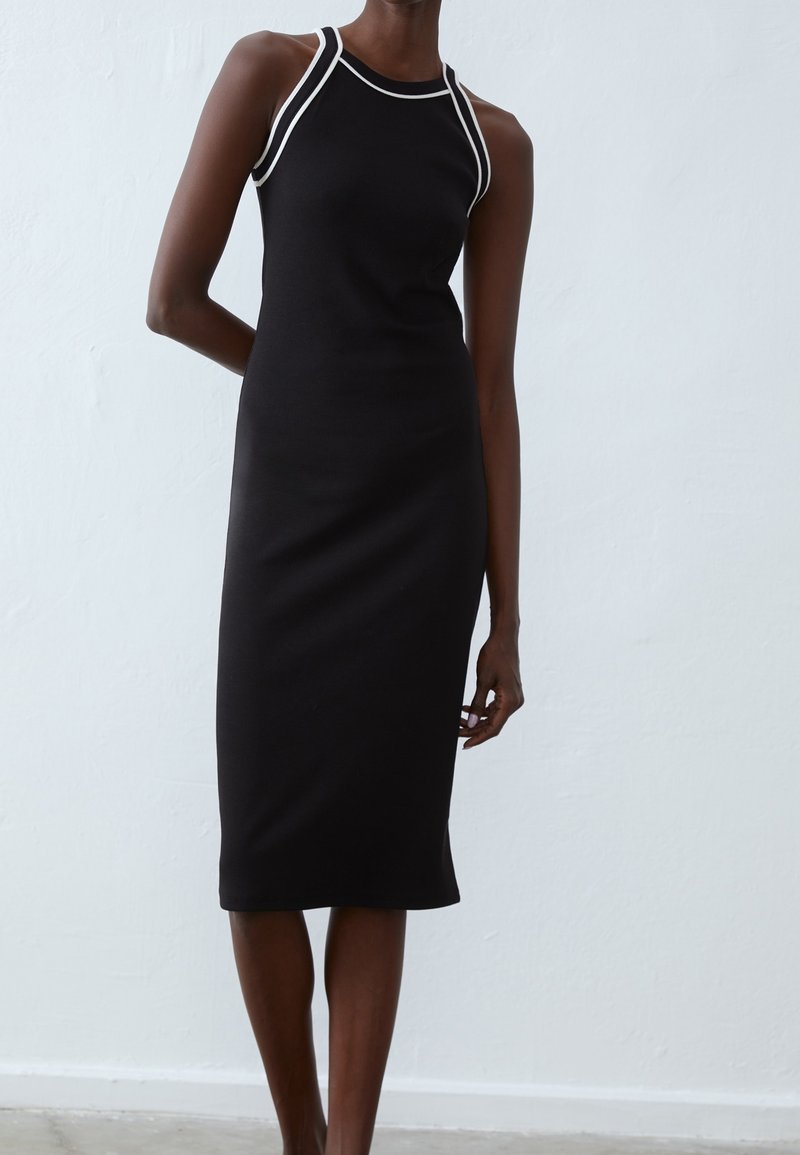 Femme portant une robe midi noire sans manches avec un liseré blanc autour du col et des emmanchures, debout devant un mur clair et uni.