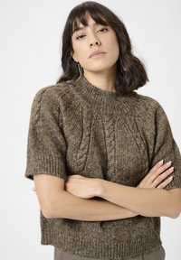 Kvinde med skulderlangt mørkt hår, iført en brun kabelstrik kortærmet sweater, med korslagte arme og lange hængende øreringe, neutral baggrund.