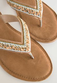 Tan flip flops met een gestructureerde suede binnenzool. De bovenkant heeft gevlochten stro en blauwe kralen, verfraaid met metallic randen.
