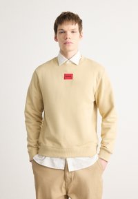 Felpa beige chiaro con collo a giro, polsini e orlo a costine, caratterizzata da un'etichetta rettangolare rossa con la scritta "HUGO" centrata sulla parte anteriore.