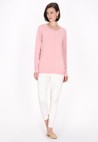 Cárdigan rosa claro con cuello redondo y botones frontales, combinado con pantalones blancos y zapatos de lona beige. Textura suave, diseño ajustado.