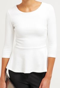 Weißes Peplum-Top mit Dreiviertelärmeln, strukturiertem Stoff und rundem Ausschnitt, kombiniert mit schwarzen Hosen, die eine taillierte Silhouette kreieren.