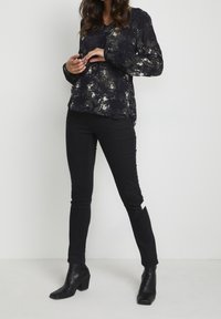 Femme se tenant avec les mains près de la taille, portant un blouse à manches longues noire à motifs, un pantalon skinny noir avec une fente à la cheville, et des bottes à talons noires.