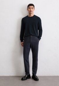 Mann steht gegen eine weiße Wand, trägt einen schwarzen langärmeligen Pullover, dunkle Navi-Hosen und schwarze Slipper, mit einer Hand in der Tasche.