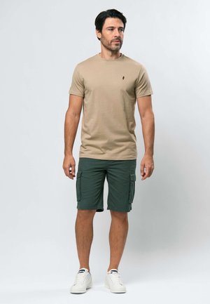 Egy férfi áll, bézs rövid ujjú pólót, sötétzöld cargo rövidnadrágot és fehér sportcipőt visel, egyszerű világos háttér előtt.