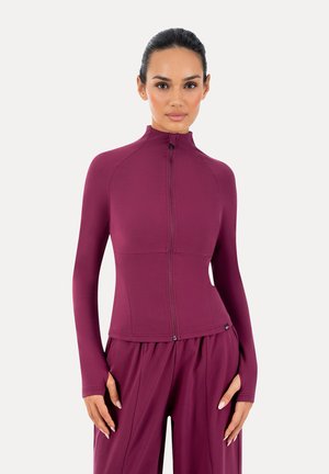 Kobieta o ciemnych włosach, ubrana w dopasowaną bluzkę na zamek z długim rękawem i luźne spodnie, oba z pasującego głębokiego fioletowego materiału, stojąca na jednolitym tle.