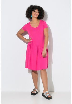 Kobieta o kręconych włosach, nosząca turkusowe kolczyki, jaskraworóżową sukienkę z krótkim rękawem oraz srebrne sandały na koturnie, stojąca na tle białej ściany.