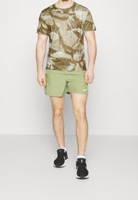 Oliivinvihreät shortsit, joissa on valkoinen Nike-logo, paritettu ruskean ja beige työpaitamaisen camo-kuosisen T-paidan kanssa. Mustat kengät, joissa on valkoiset pohjat.