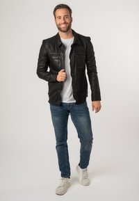 Giubbotto in pelle nera con tasche frontali, indossato sopra una camicia bianca, abbinato a jeans blu e sneakers bianche. Texture liscia, design aderente.